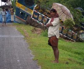 An over-turned bus leaves passengers on the side of a road.